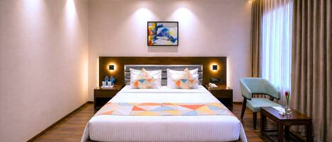 A hotel room with a large bed, a headboard with integrated lighting, a bedside table with a phone, a chair, and a window with curtains.