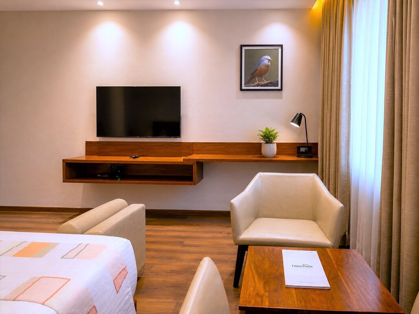 A hotel room with a flat-screen TV, a wooden shelf with a plant, a desk with a notebook, and two chairs.