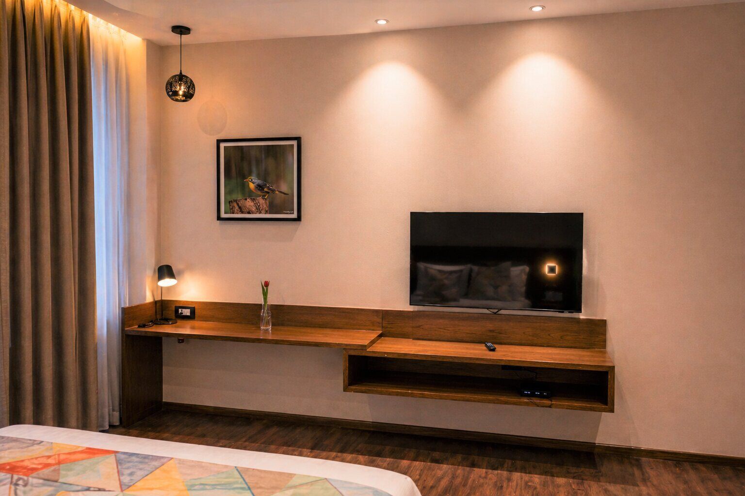 A modern hotel room with a flat-screen TV mounted on the wall, a wooden console table, a bedside lamp, and a framed picture above the console.