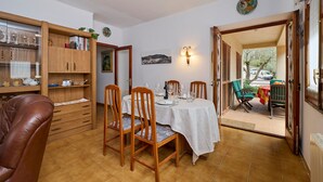 Een eetruimte met een tafel voor vier personen, houten stoelen, een kast met keukengerei en uitzicht door een open deur op een buitenruimte.