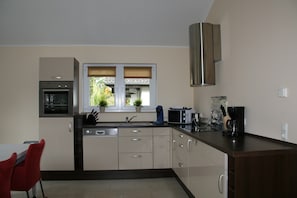 A modern kitchen with a dishwasher, oven, microwave, and a window with blinds. - Bungalow in Eifel Near Forest and Mountains (Gerolstein)