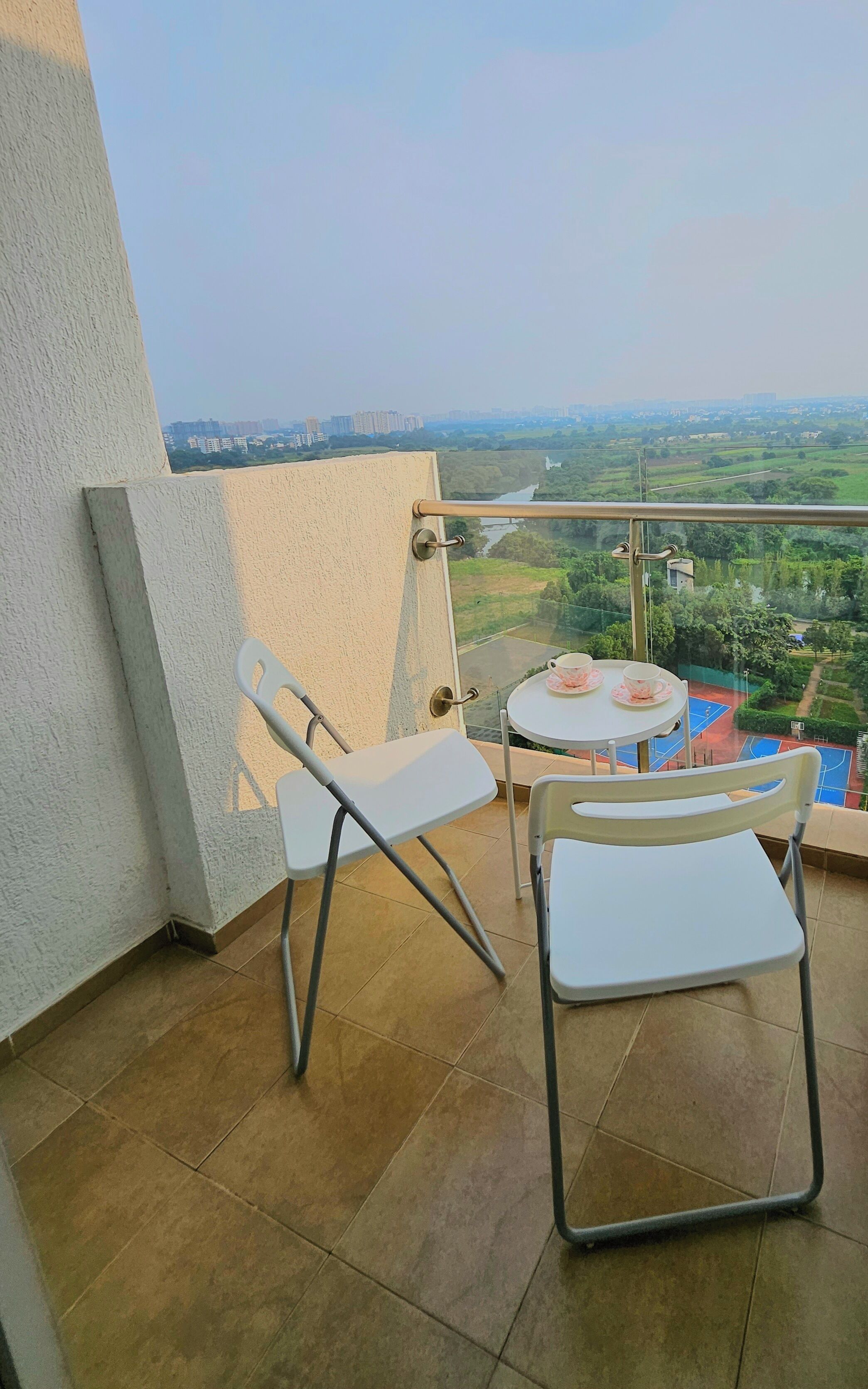 A balcony with two white chairs, a small round table with a cup and saucer, and a view of a sports court and buildings in the distance.