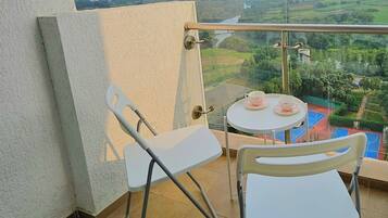A balcony with two white chairs, a small round table with a cup and saucer, and a view of a sports court and buildings in the distance.