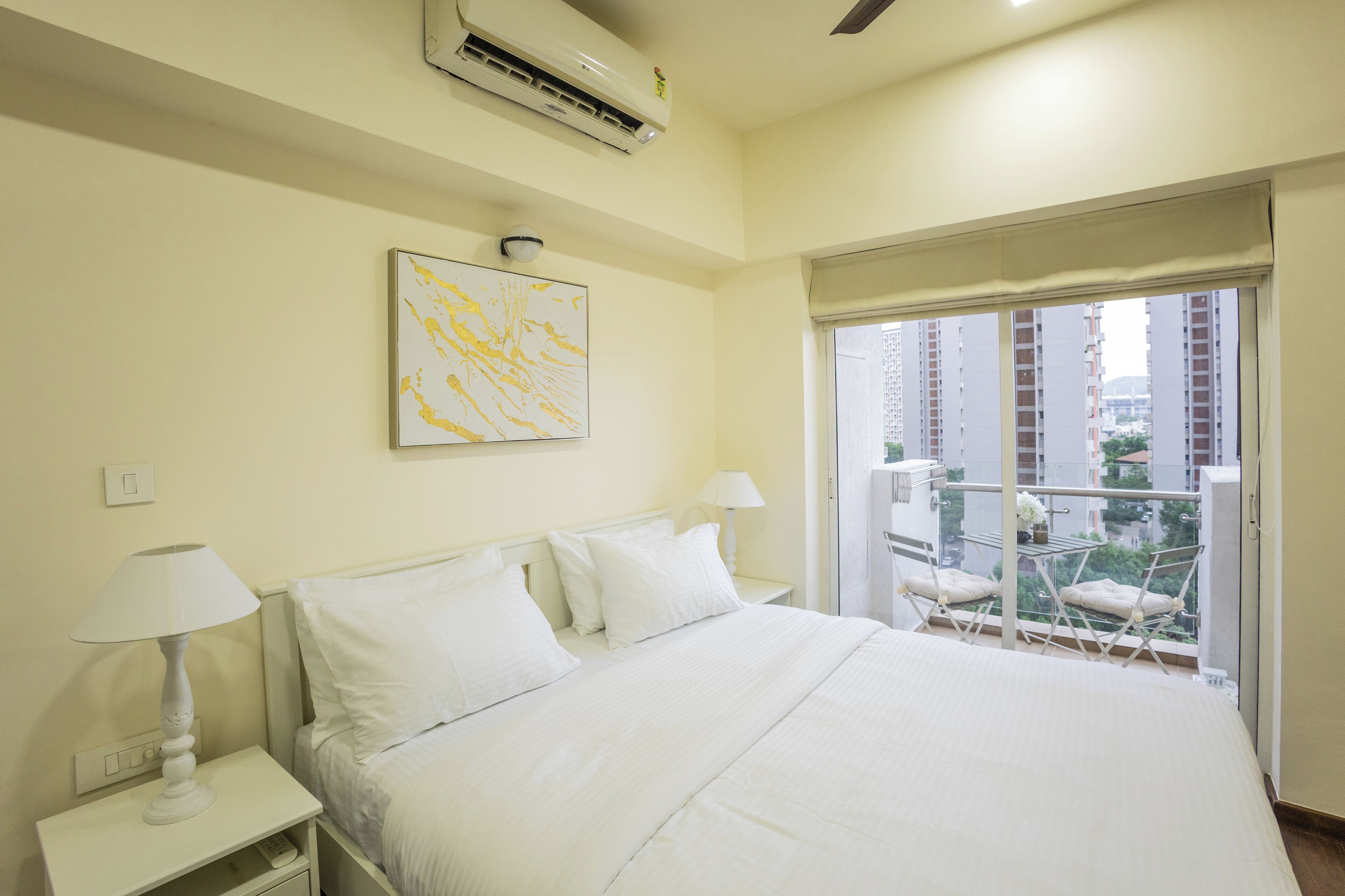 Ein Hotelzimmer mit Bett, weißen Nachttischen, einer Wand-Klimaanlage, einem Deckenventilator, einem Fenster mit Blick auf Gebäude und einem Balkon mit Stühlen.