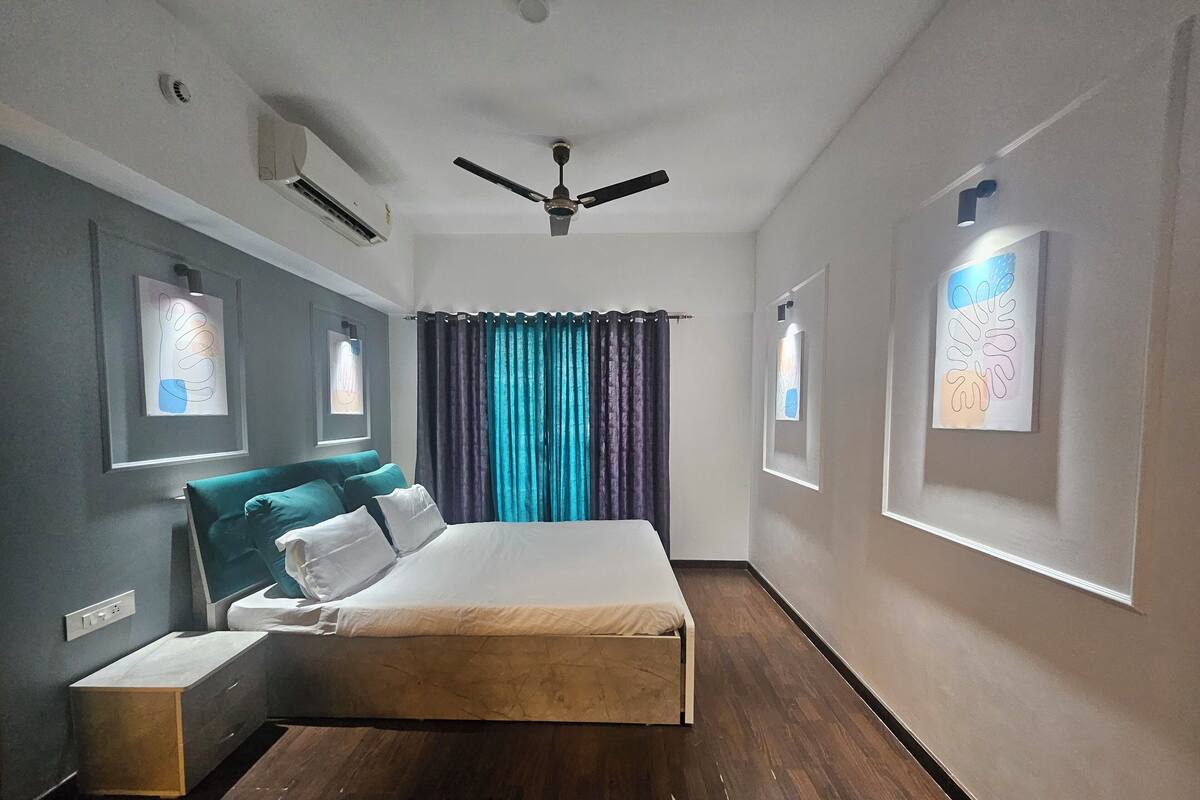 A hotel room with a bed, a ceiling fan, air conditioning, a window with curtains, and artwork on the walls.