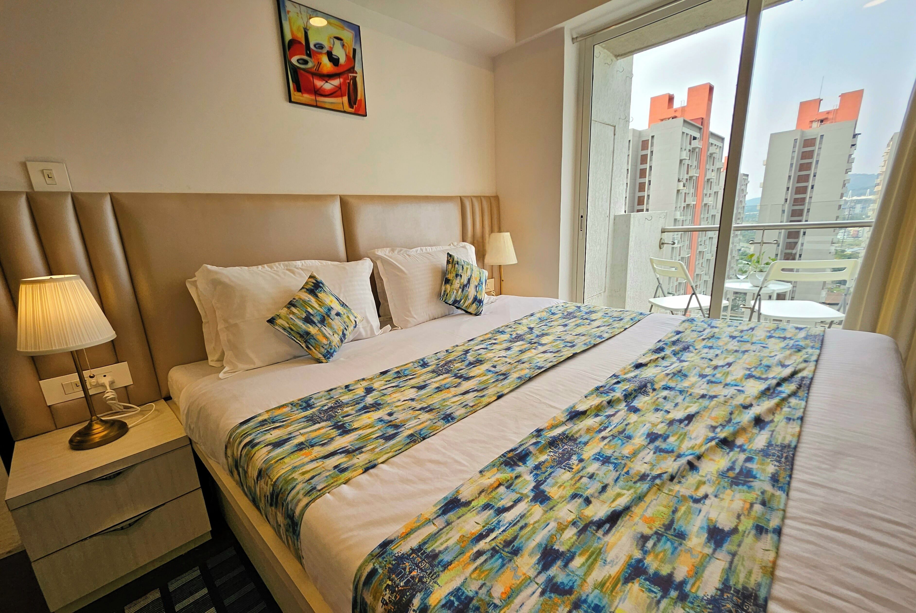 Hotellihuone, jossa on sänky, yöpöytä, lamppu ja ikkunasta avautuva näkymä rakennuksiin.