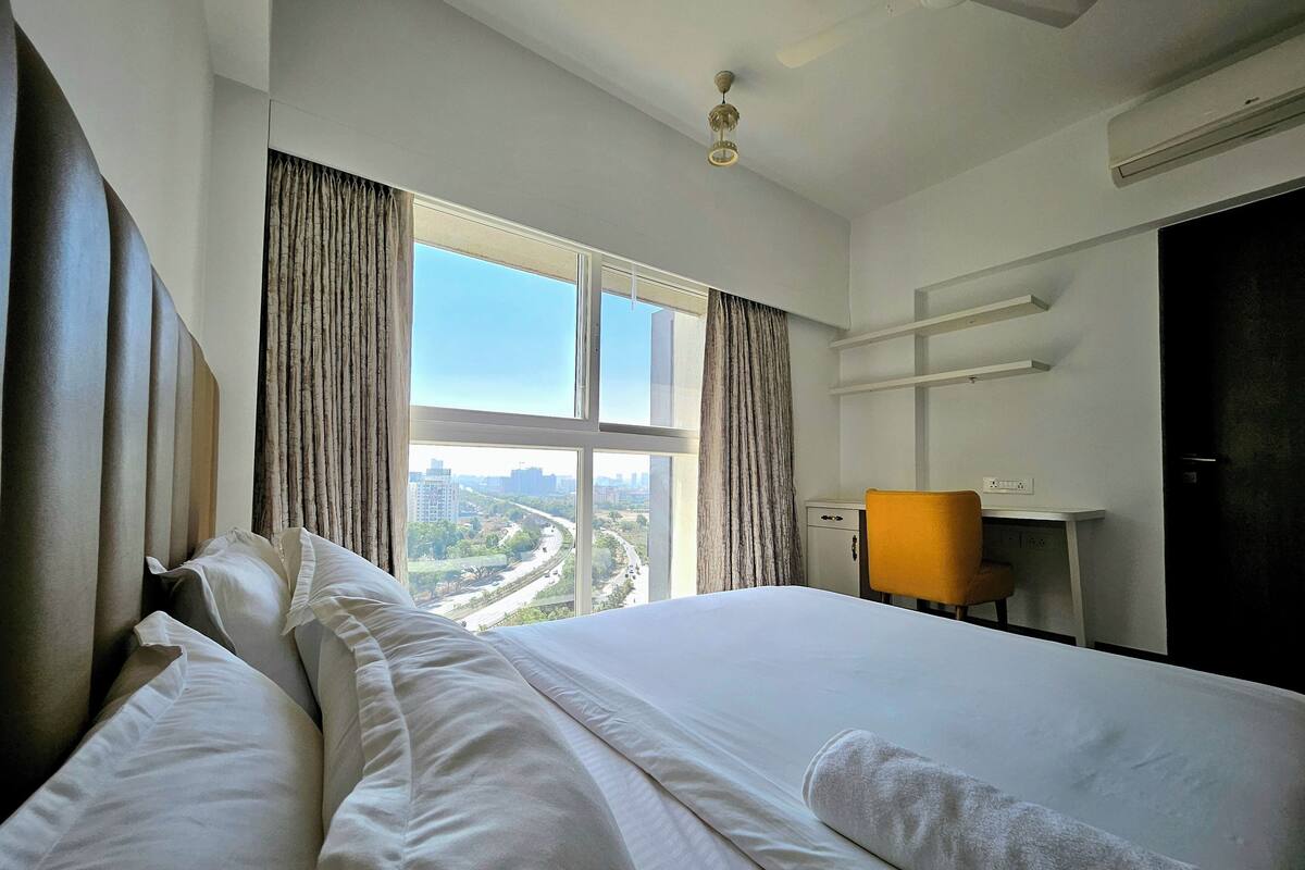 A hotel room with a large bed, a desk, a chair, and a view of a cityscape through the window.