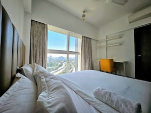 A hotel room with a large bed, a desk, a chair, and a view of a cityscape through the window.