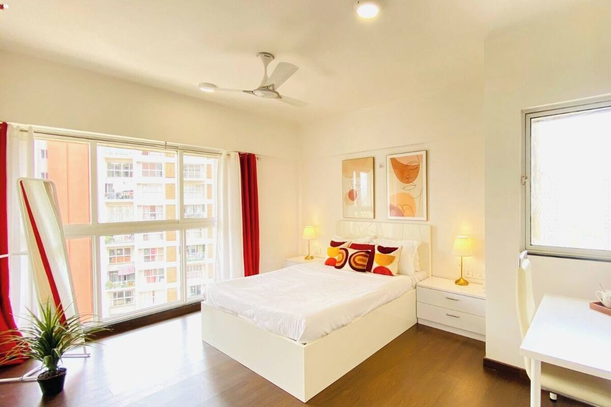 A bedroom with a bed, bedside tables, a desk, a ceiling fan, and a window with a view of buildings.