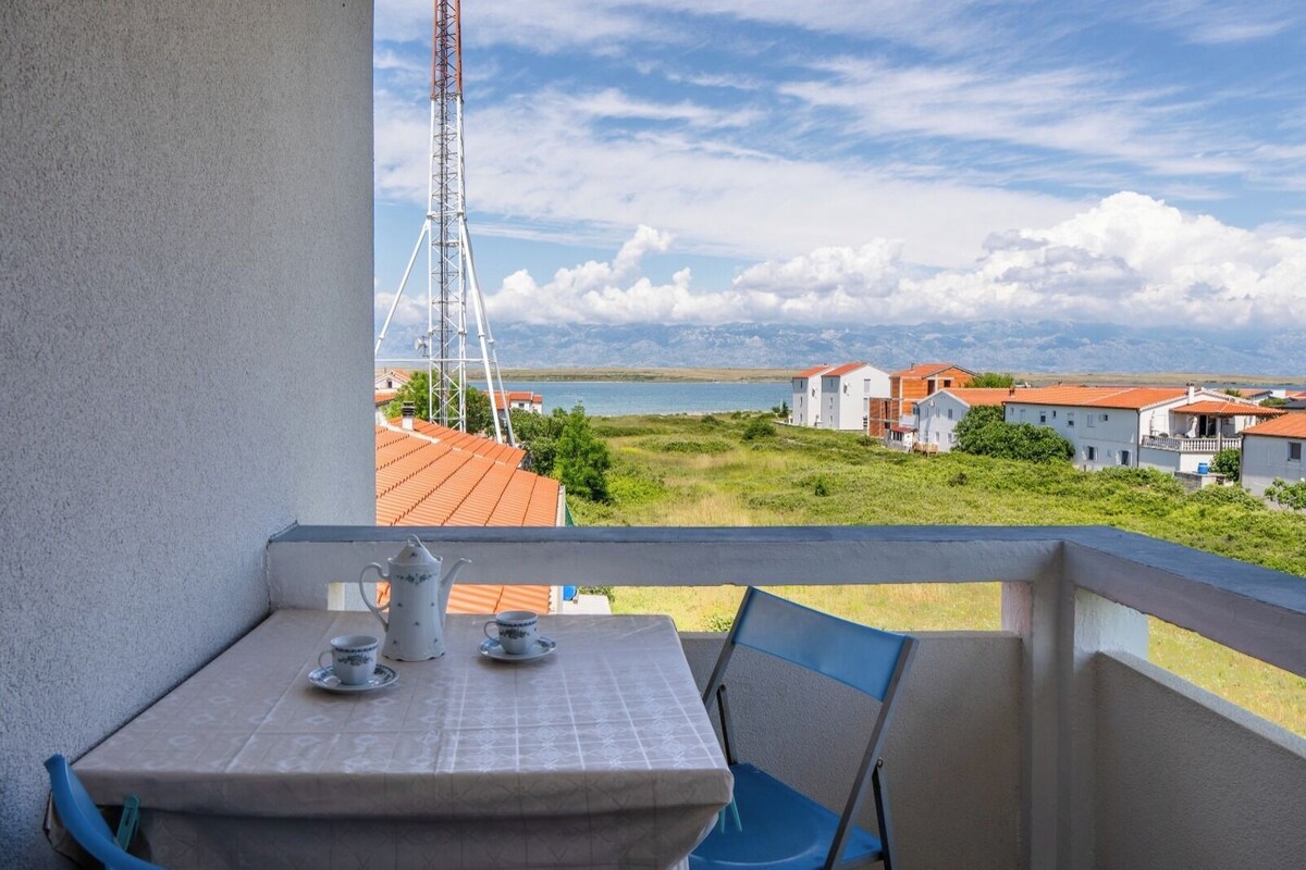 Ein Balkon mit Tisch für zwei Personen, mit Blick auf eine Küstenlandschaft mit Gebäuden und wolkenlosem Himmel.