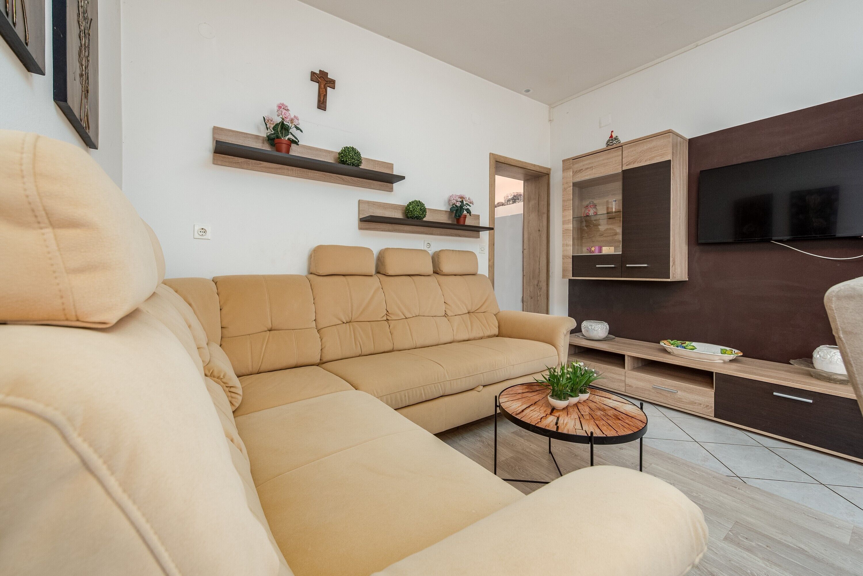 A living room with a beige sofa, a coffee table with a plant, wooden shelves with flowers, a TV, and a cabinet.