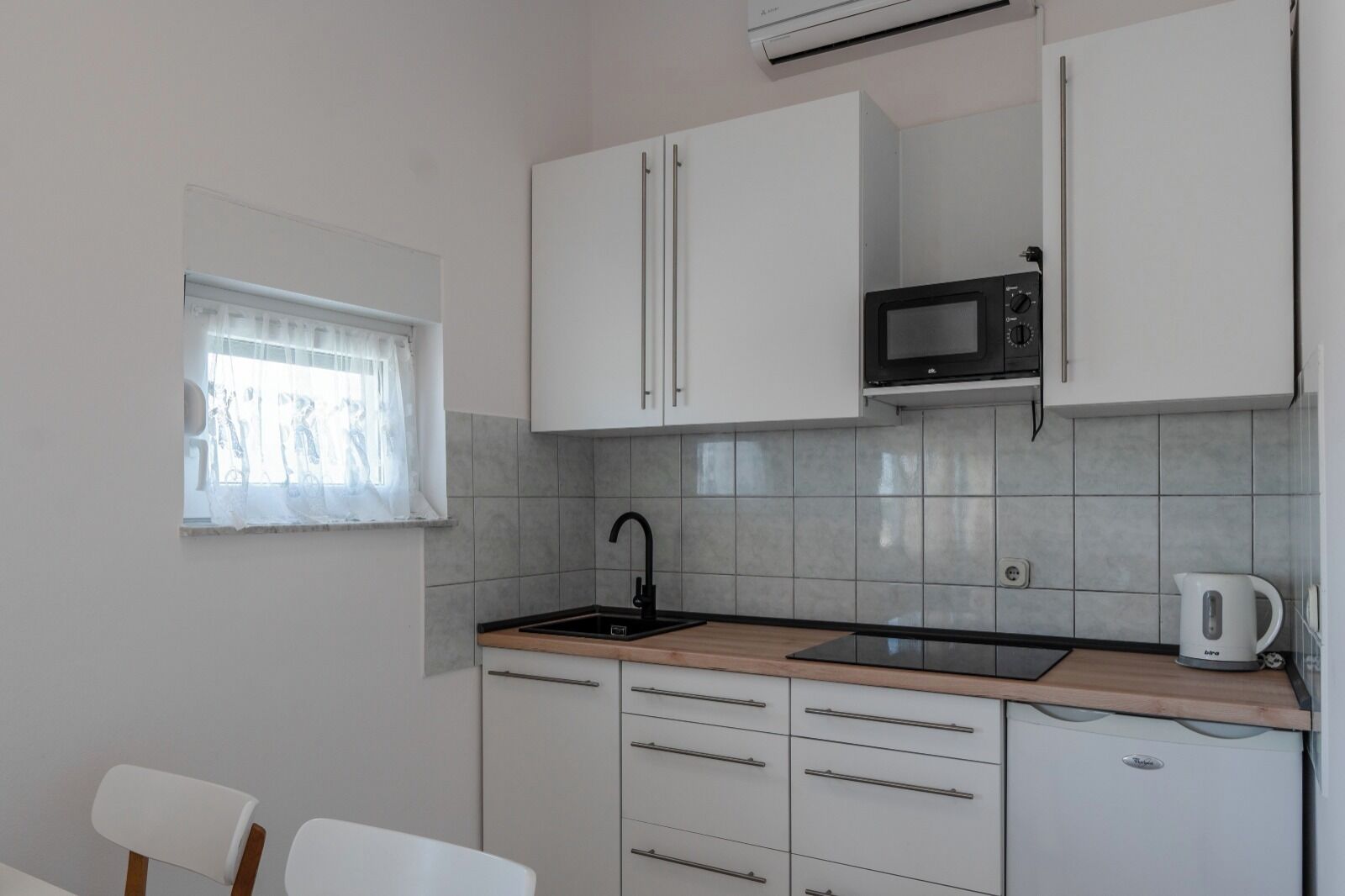 A compact kitchen with white cabinets, a black sink, a microwave, and a kettle.