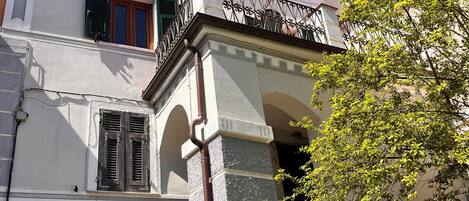 Un bâtiment à la façade blanche, une porte noire ornée d’un motif géométrique et un balcon avec un garde-corps en fer forgé.