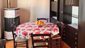 Een eetruimte met een ronde tafel, overdekt met een tafelkleed met bloemenprint, houten stoelen, een fornuis, een klok aan de muur en een kast met glazen deuren.