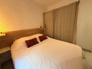 A neatly made bed with white bedding and two brown pillows, a wooden headboard, a bedside table with a lamp, and a window with beige curtains. - Apartment with AC, parking - 4 guests (Banyuls-sur-Mer)