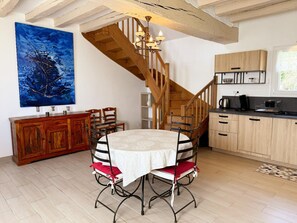 A dining area with a round table, chairs, a wooden staircase, and a kitchen in the background. - Authentic gîte with garden - 4 guests (Thésée)
