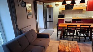 Een woonkamer met een bank en een houten tafel, een keuken met een koelkast, magnetron, fornuis en gootsteen, en een eethoek met stoelen en een tafel.