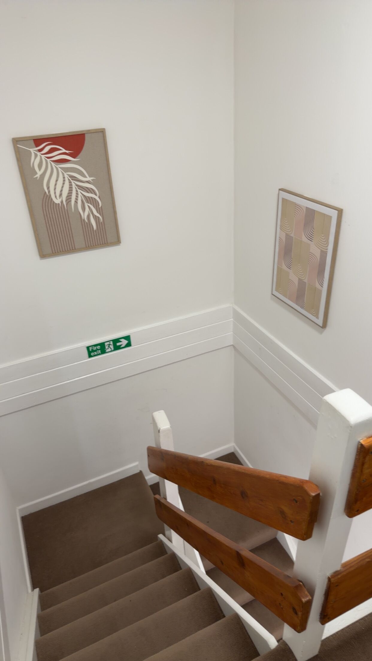 A staircase with wooden handrails, a fire exit sign, and two framed artworks on the walls.