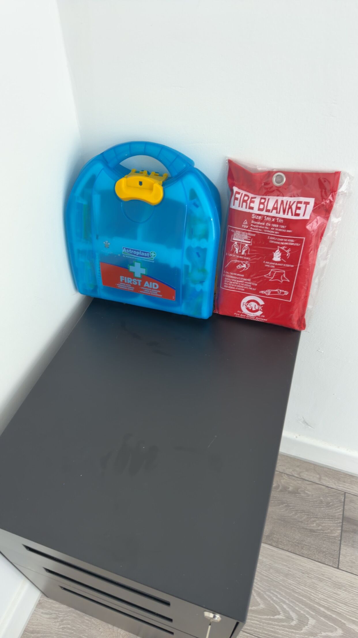 Una caja de almacenamiento negra con un extintor azul y una manta ignífuga roja encima.
