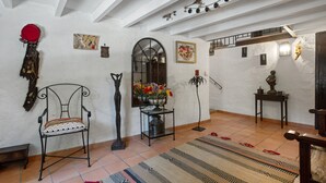 Une pièce avec un plafond blanc, une chaise dotée d’un coussin à motifs, une table sur laquelle est posé un vase de fleurs et un tapis sur le sol.