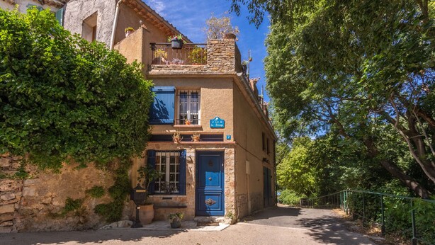 Un bâtiment traditionnel avec une façade en pierre, une porte bleue et un balcon agrémenté de plantes en pot.