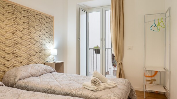 Une chambre d’hôtel avec une tête de lit à motifs, un lit recouvert d’une literie à fleurs, une serviette soigneusement pliée, une petite table de chevet avec une lampe et un balcon doté d’une balustrade.