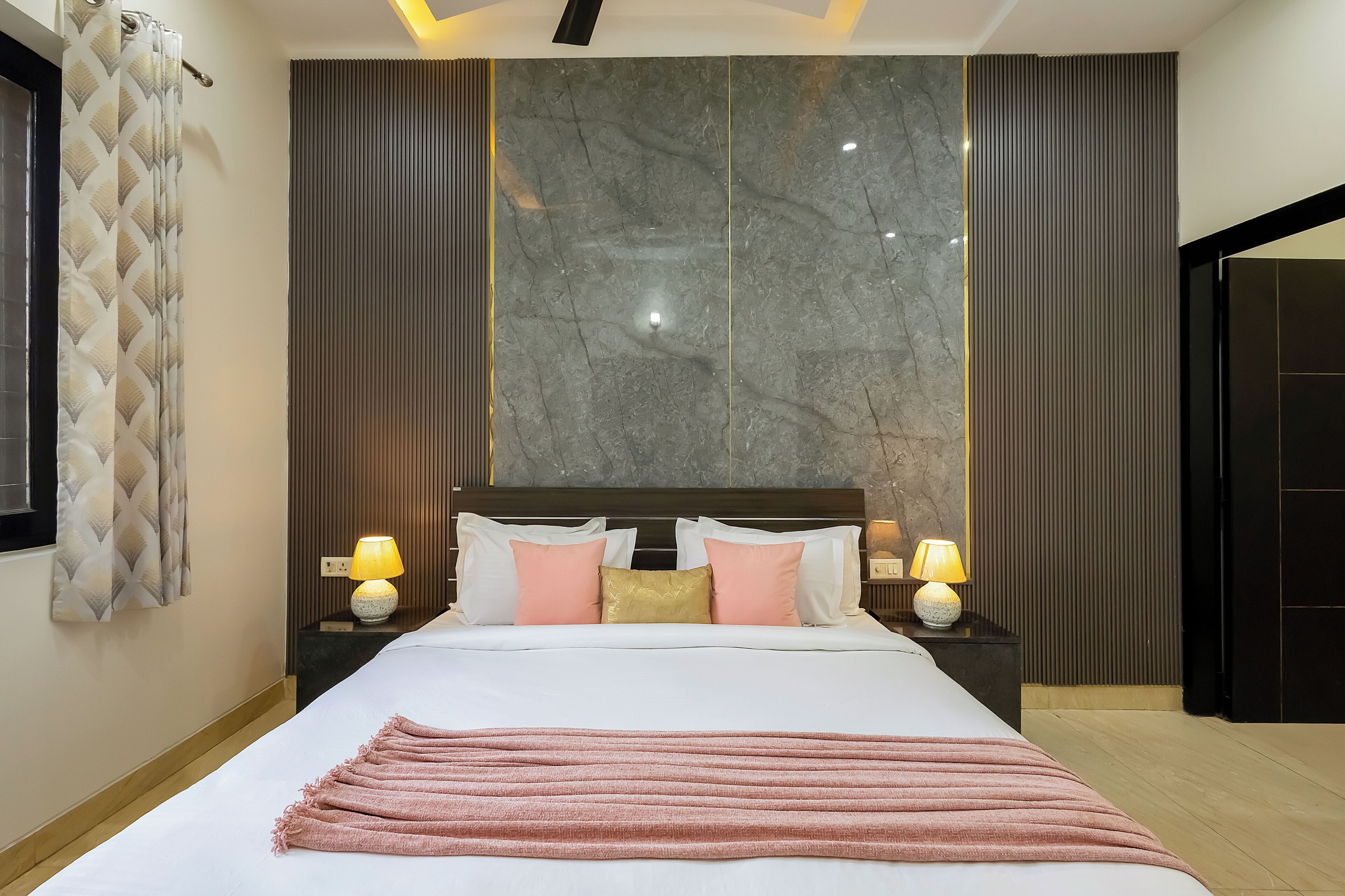 Ein modernes Hotelzimmer mit einem großen Bett, zwei Nachttischlampen, einer Marmorwand und einem gemusterten Vorhang.