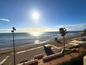 Blick auf einen Strand mit blauem Himmel, der Sonne, die sich im Wasser spiegelt, und einem Rettungsturm.