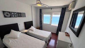 Ein Hotelzimmer mit zwei Betten, einem Schreibtisch, einem Stuhl, einem Deckenventilator, einem Fenster mit Meerblick und einer Wand-Klimaanlage.