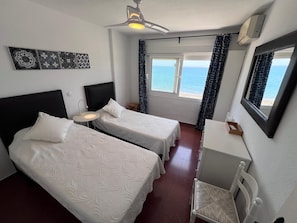 Ein Hotelzimmer mit zwei Betten, einem Schreibtisch, einem Stuhl, einem Deckenventilator, einem Fenster mit Meerblick und einer Wand-Klimaanlage.