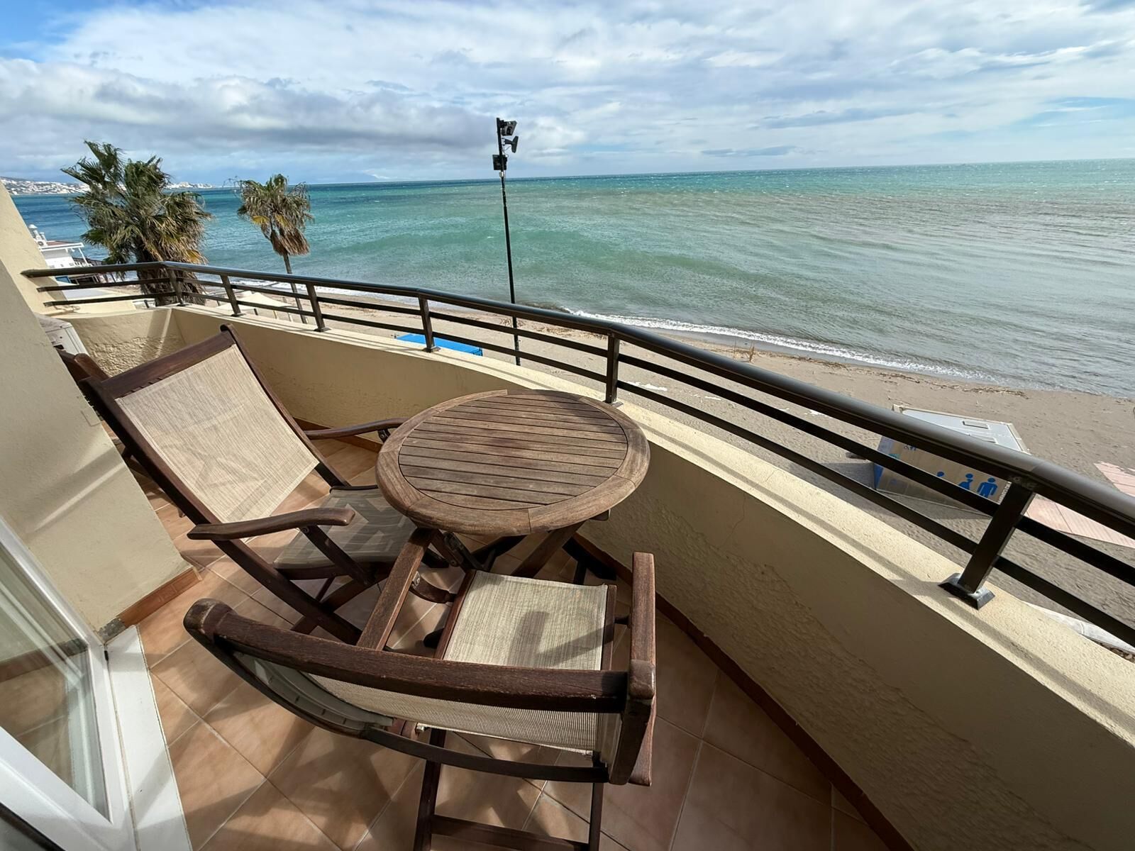 A balcony with a wooden table and two chairs, overlooking a beach with clear water and palm trees.