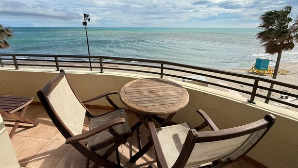 Ein Balkon mit rundem Holztisch und Stühlen, Blick auf den Strand und einem Rettungsschwimmerstand.
