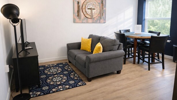 A living room with a gray sofa, yellow pillows, a black floor lamp, a patterned rug, a wooden dining table with chairs, and a large window with blinds. - Downtown Detroit Swanky Suite (Detroit)