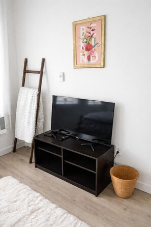 Una habitación con una televisión de pantalla plana sobre un soporte de madera oscura, una toalla blanca colgada en una escalera de madera, una cesta de mimbre y una obra de arte enmarcada en la pared.
