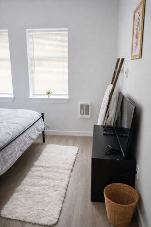 Een slaapkamer met een bed, een witte vloerkleed, een zwarte tv-kast en een raam met luxaflex.