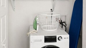 A front-loading washing machine with a control panel, a white laundry basket, an iron, and folded blankets on a shelf above. - Downtown Detroit Swanky Suite (Detroit)