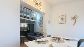 Comedor con una mesa blanca dispuesta para cuatro, cocina al fondo y balcón con barandilla.
