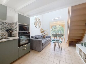 Ein kompakter Wohnbereich mit Küchenzeile, einem grauen Sofa, einem Esstisch und einer Treppe.