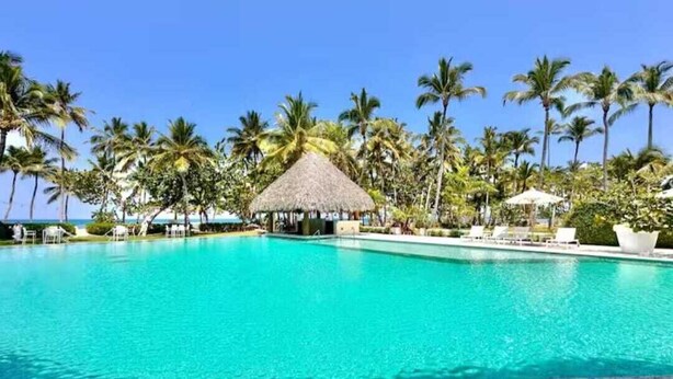 A clear turquoise pool surrounded by palm trees and a thatched-roof structure. - Cozy studio at Portillo, Las Terrenas (Las Terrenas. El Portillo Residences.)
