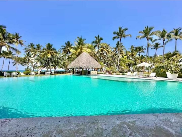 A clear turquoise pool surrounded by palm trees and a thatched-roof structure. - Cozy studio at Portillo, Las Terrenas (Las Terrenas. El Portillo Residences.)
