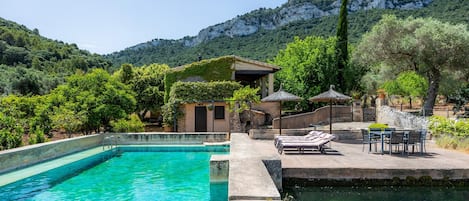 A swimming pool with lounge chairs and a table, surrounded by greenery and a mountain backdrop.