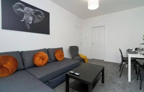 Ein Wohnzimmer mit grauem Sofa, orangenen Kissen, einem schwarzen Couchtisch, einem Esstisch mit Stühlen und einem großen Elefantenbild an der Wand.