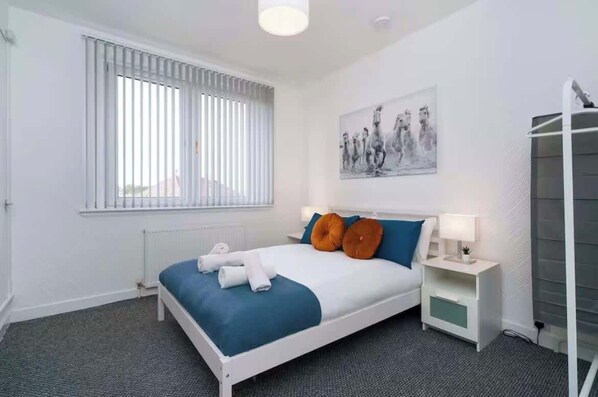 Ein Schlafzimmer mit Bett, Fenster mit Jalousien, einem Bild an der Wand und einem Nachttisch mit Lampe.