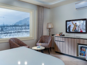A hotel room with a large window, a television, a dining table, two patterned armchairs, a lamp, and a minibar. - Faiz Resort Pahalgam (Pahalgam)