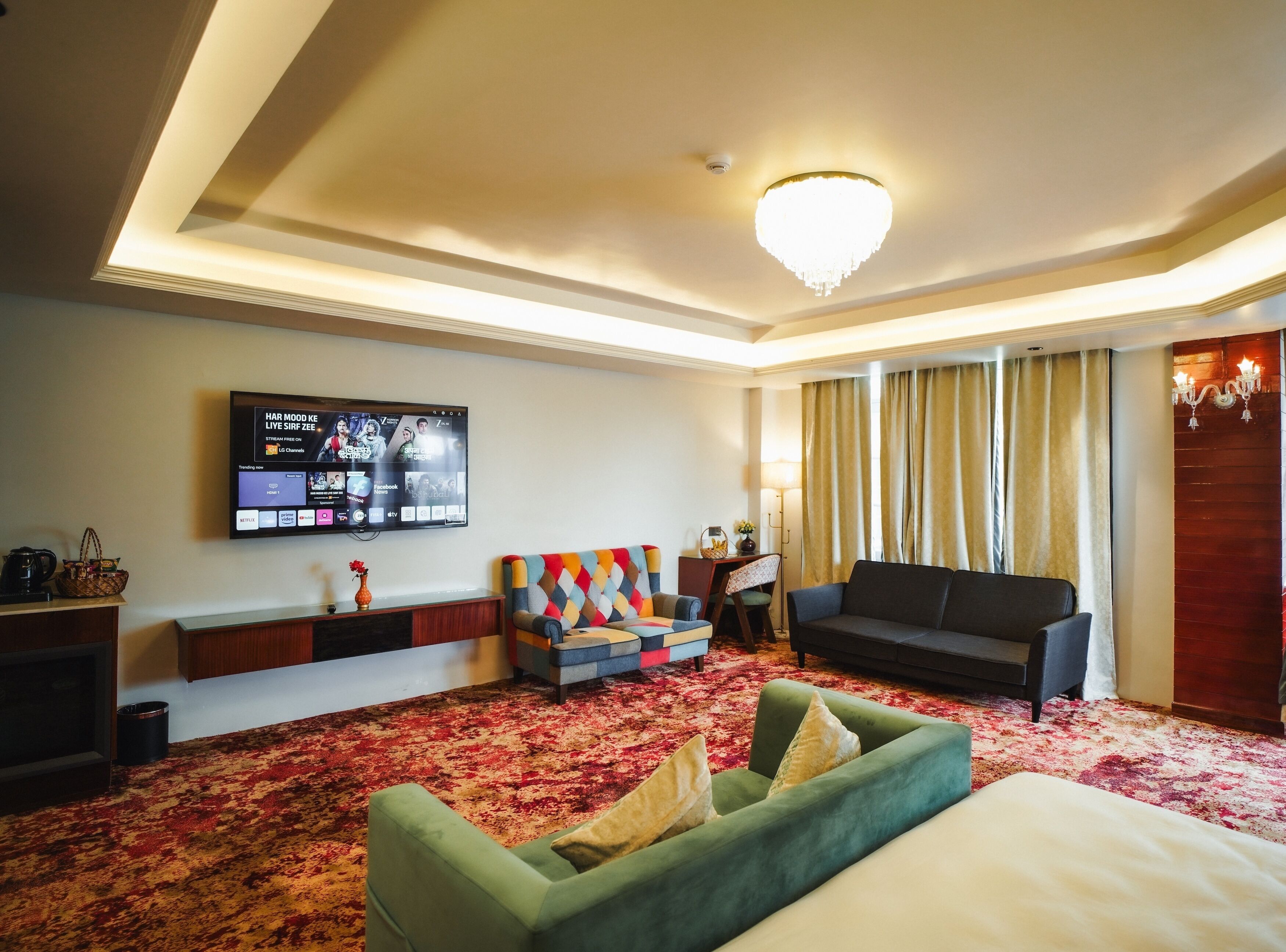 Habitación de hotel con una gran televisión de pantalla plana, un sofá verde, un sofá negro, un sillón estampado, una alfombra roja y beige, y una araña.