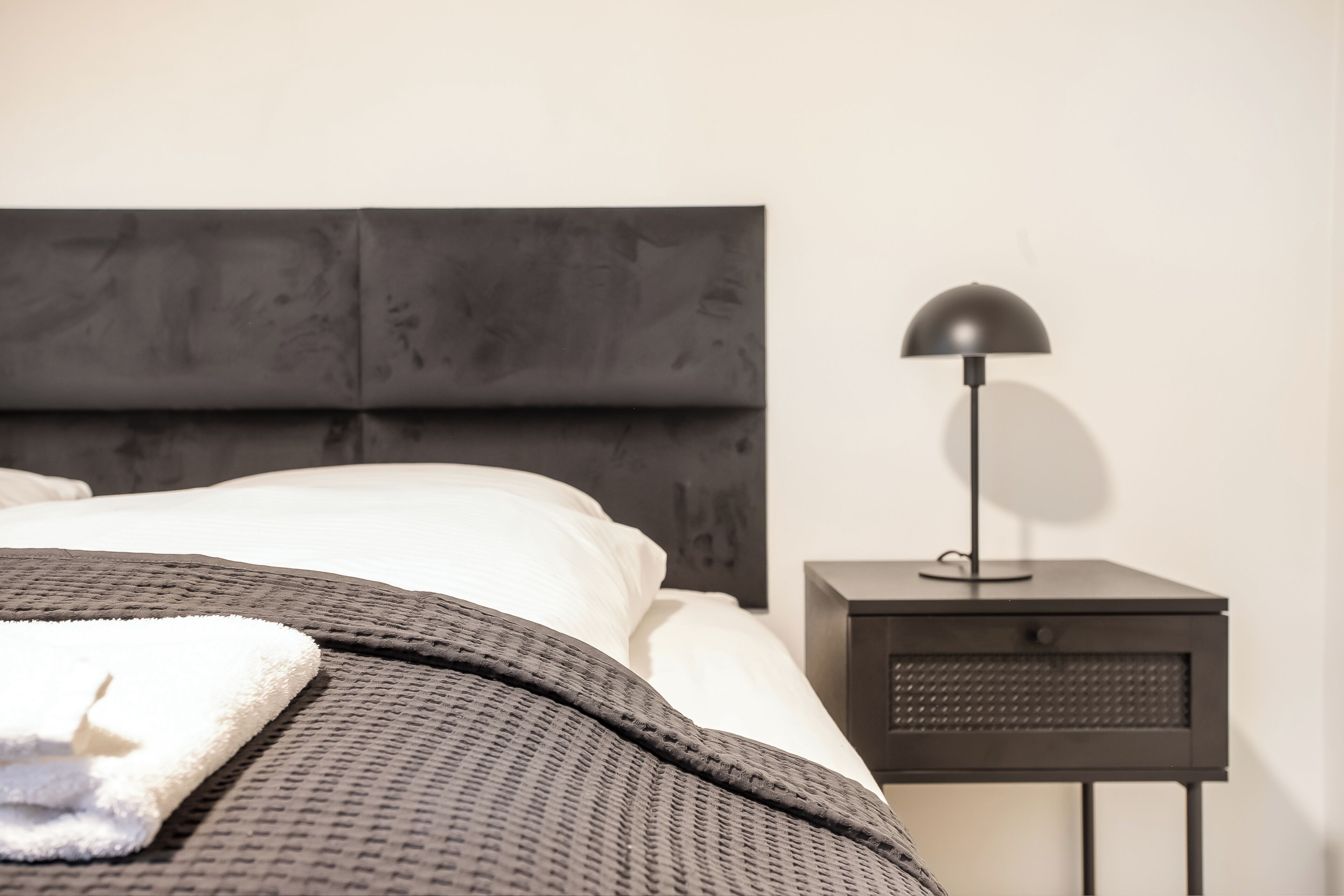 Una cama con cabecera oscura, almohada blanca y manta gris. Se observa una mesita de noche con una lámpara y un cajón.
