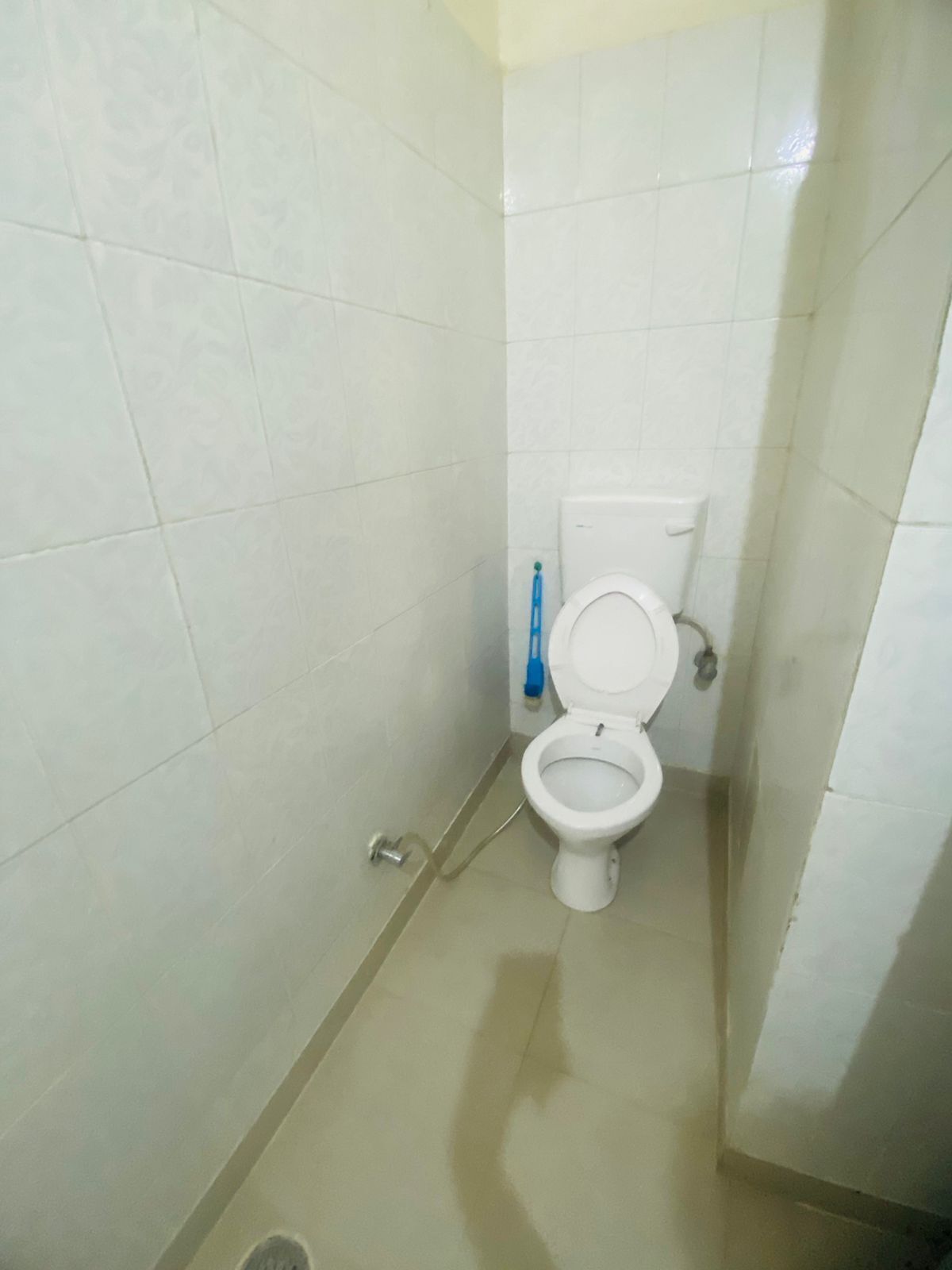 A small, clean bathroom with white tiles, a toilet, a blue cleaning brush, and a metal drain cover.