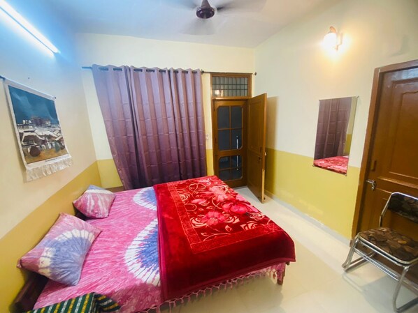 Un dormitorio con cama, cortinas, una puerta, un taburete pequeño y una lámpara fijada a la pared.