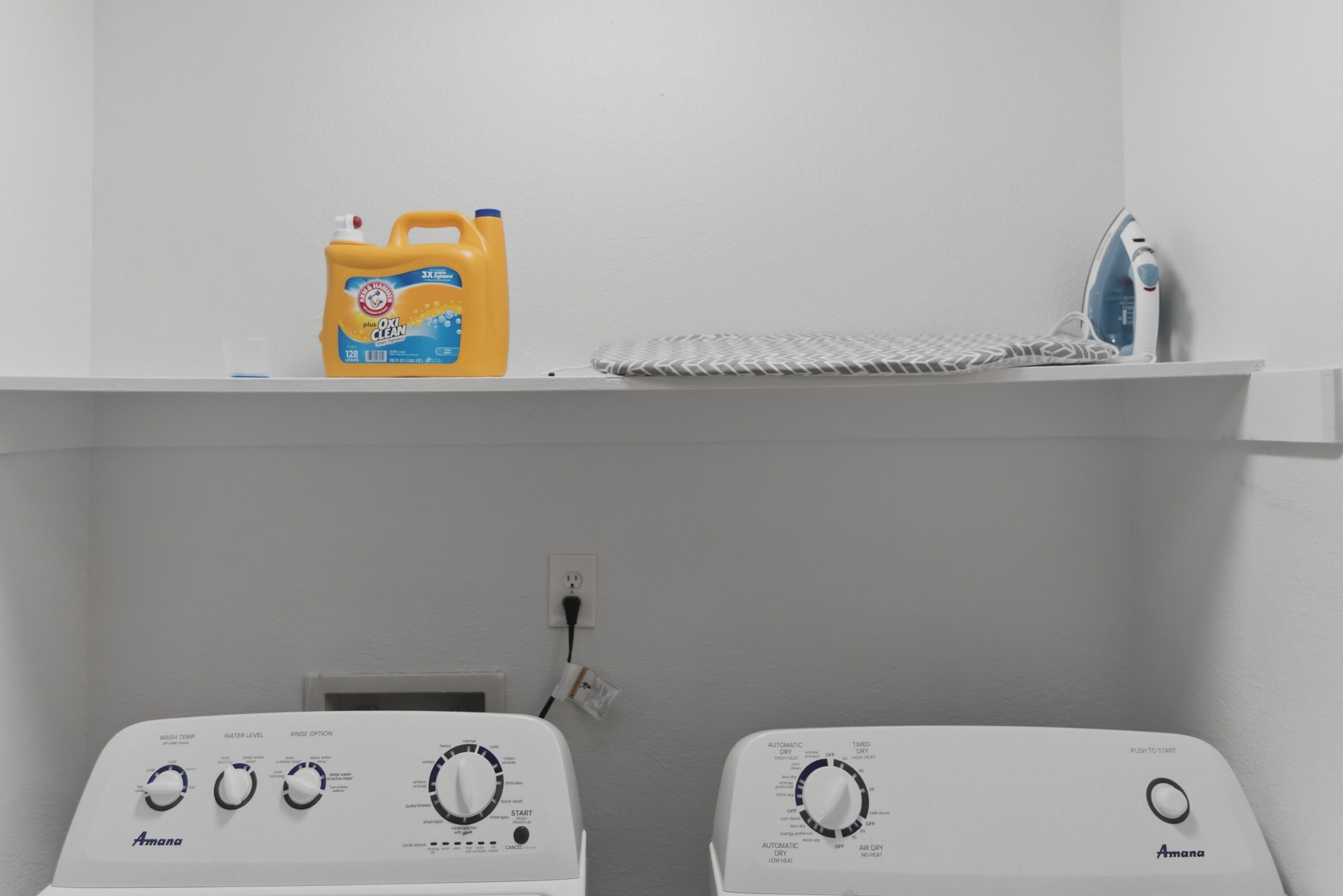 Une buanderie équipée de deux machines à laver Amana, d’une bouteille de détachant OxiClean et d’un fer à repasser posé sur une étagère.