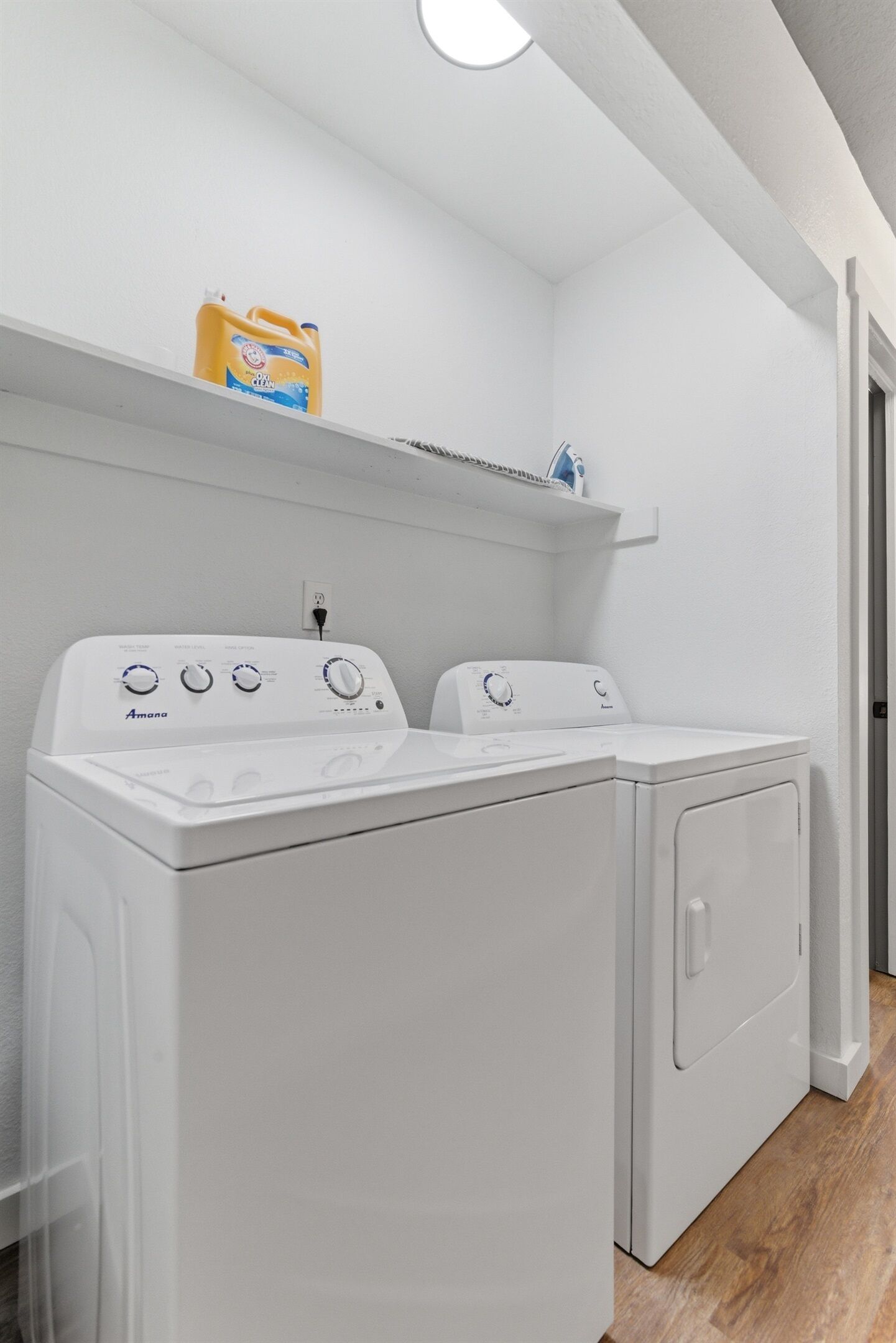 A laundry room with two white front-loading washing machines, a shelf with detergent, and a door leading to another room.